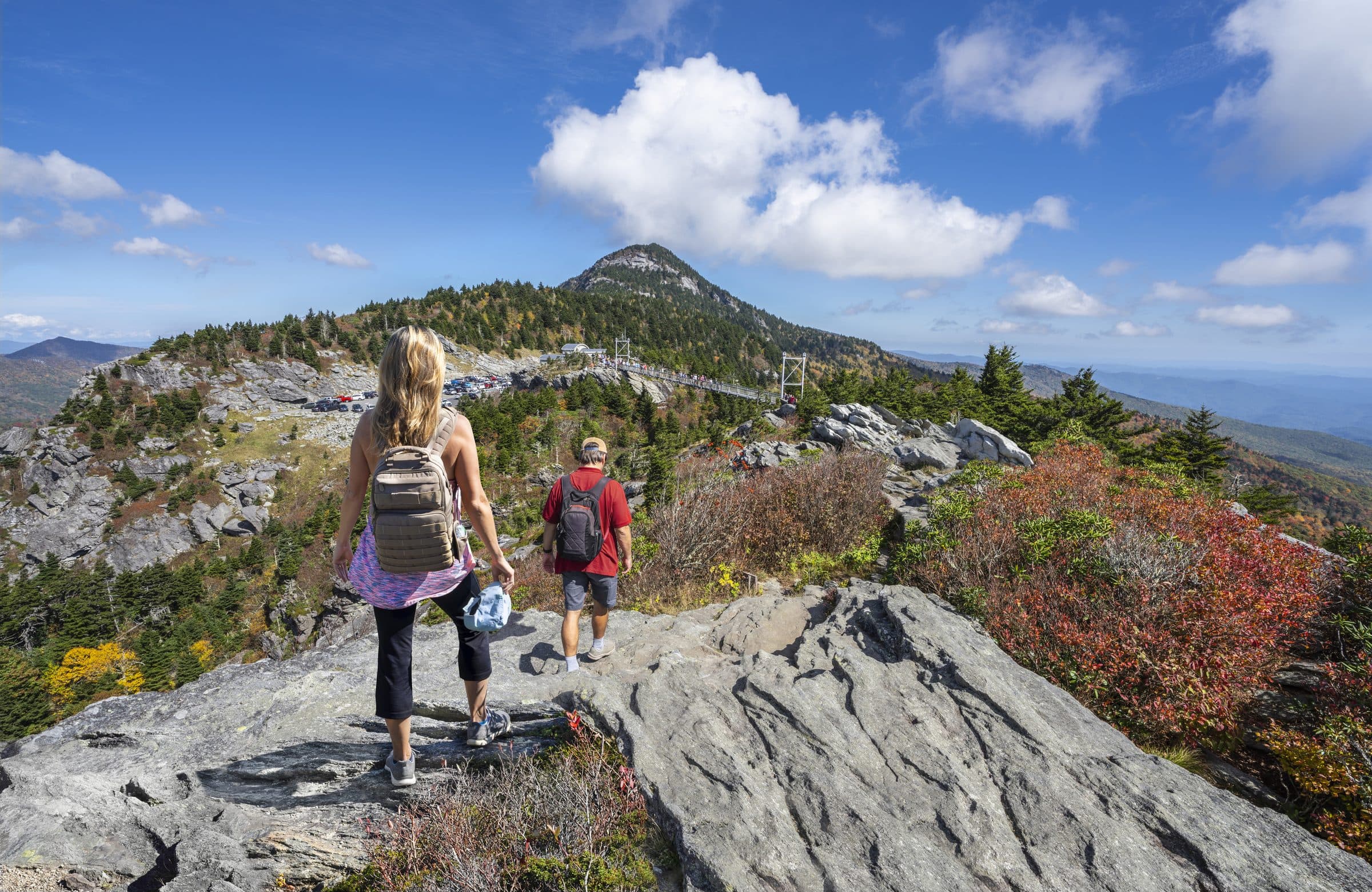 Two hikers on a rocky Grandfather Mountain trail in fall color, with the Mile High Swinging Bridge and peak behind