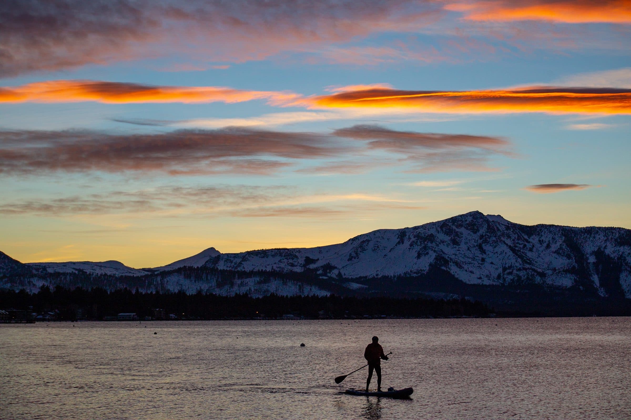 Paddleboarder silhouetted at sunset on Lake Tahoe, snowy peaks behind