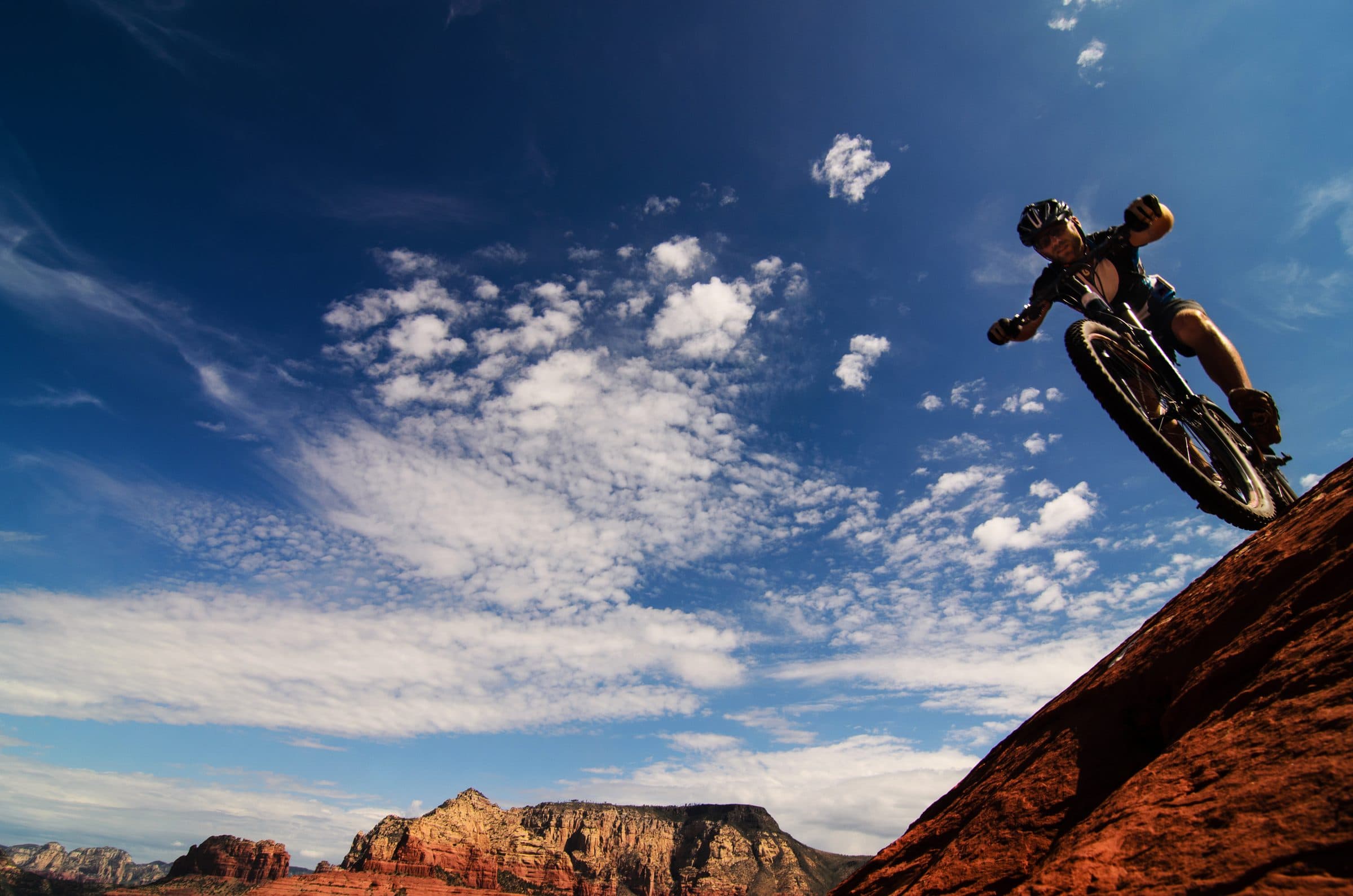 Mountain biker riding down red slickrock under a big blue sky, red buttes behind