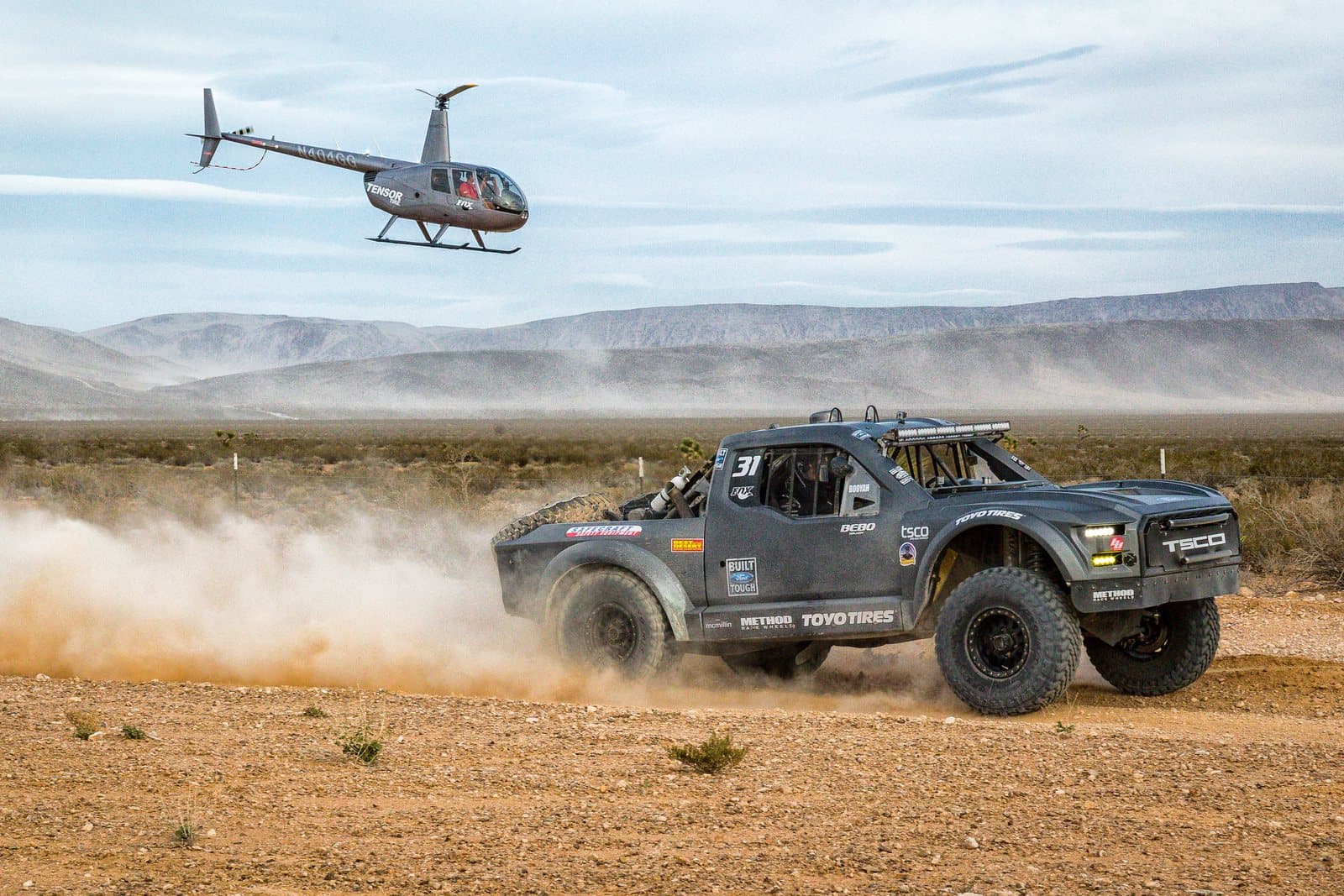 A desert race truck kicking up a dust plume with a helicopter flying alongside