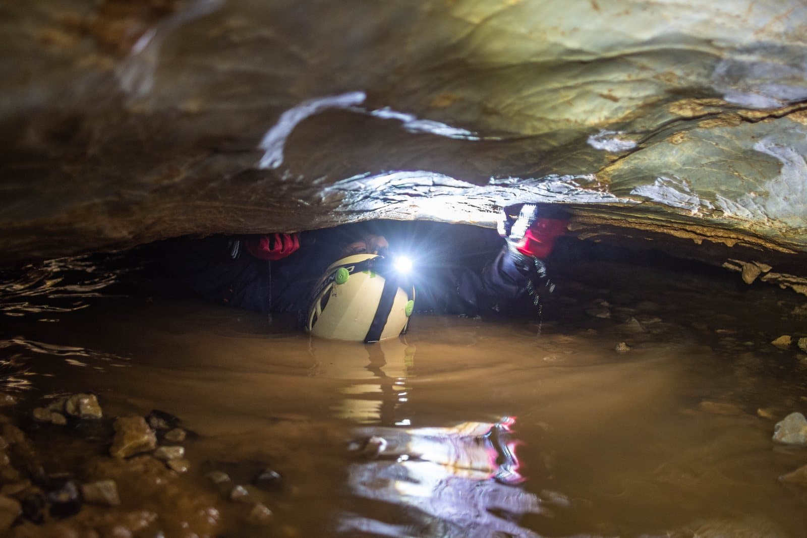 A caver with a headlamp crawling on his back through a low, flooded cave passage