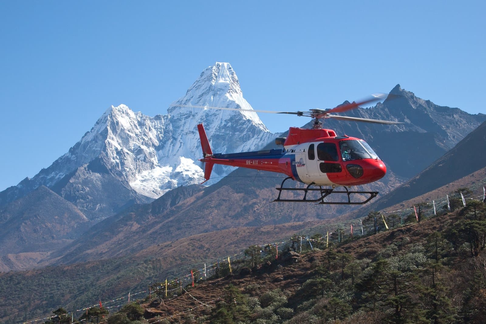 A red and white helicopter flying past snowy Himalayan peaks near Tengboche, Nepal