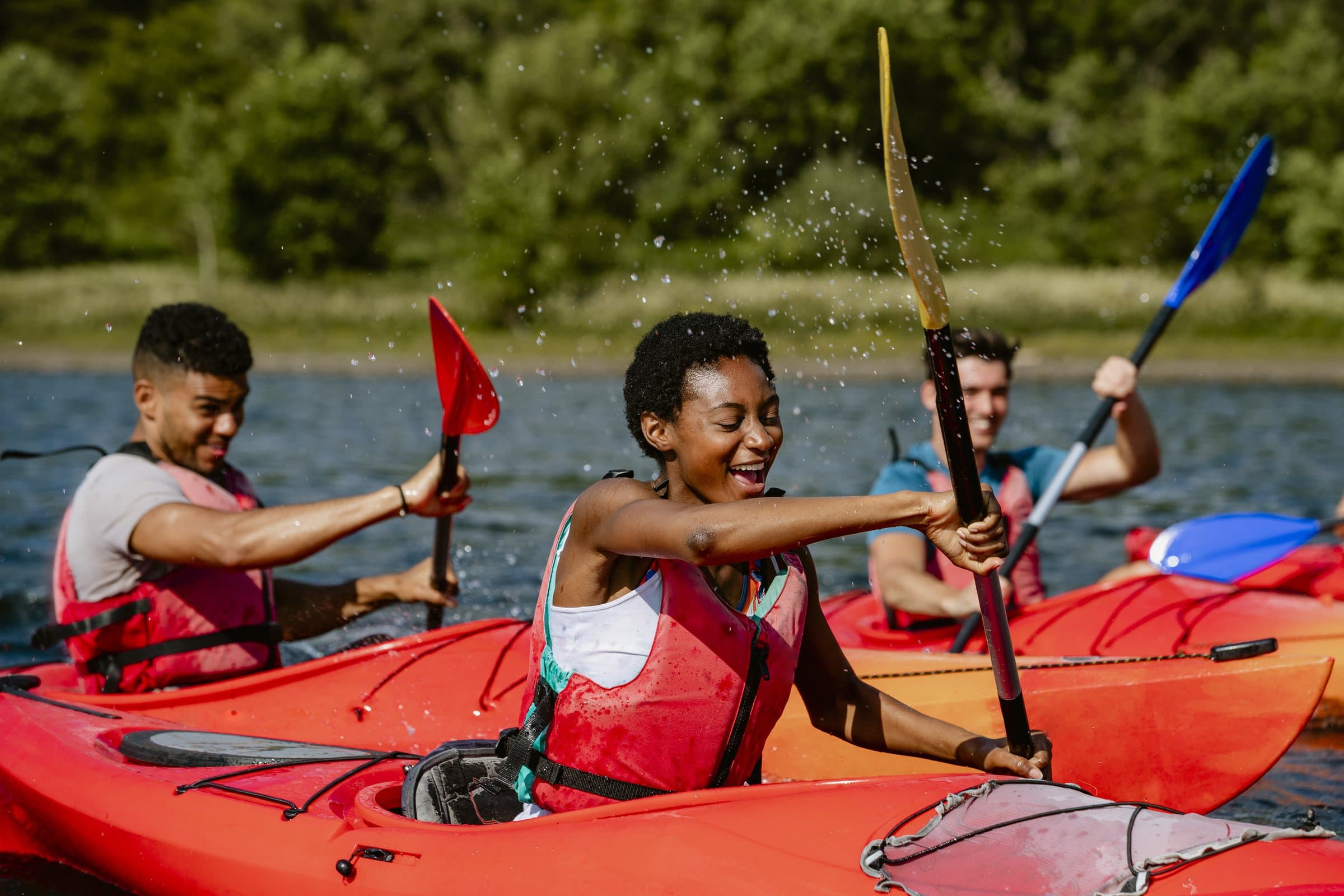 Three friends laughing and paddling red kayaks on a sunny lake