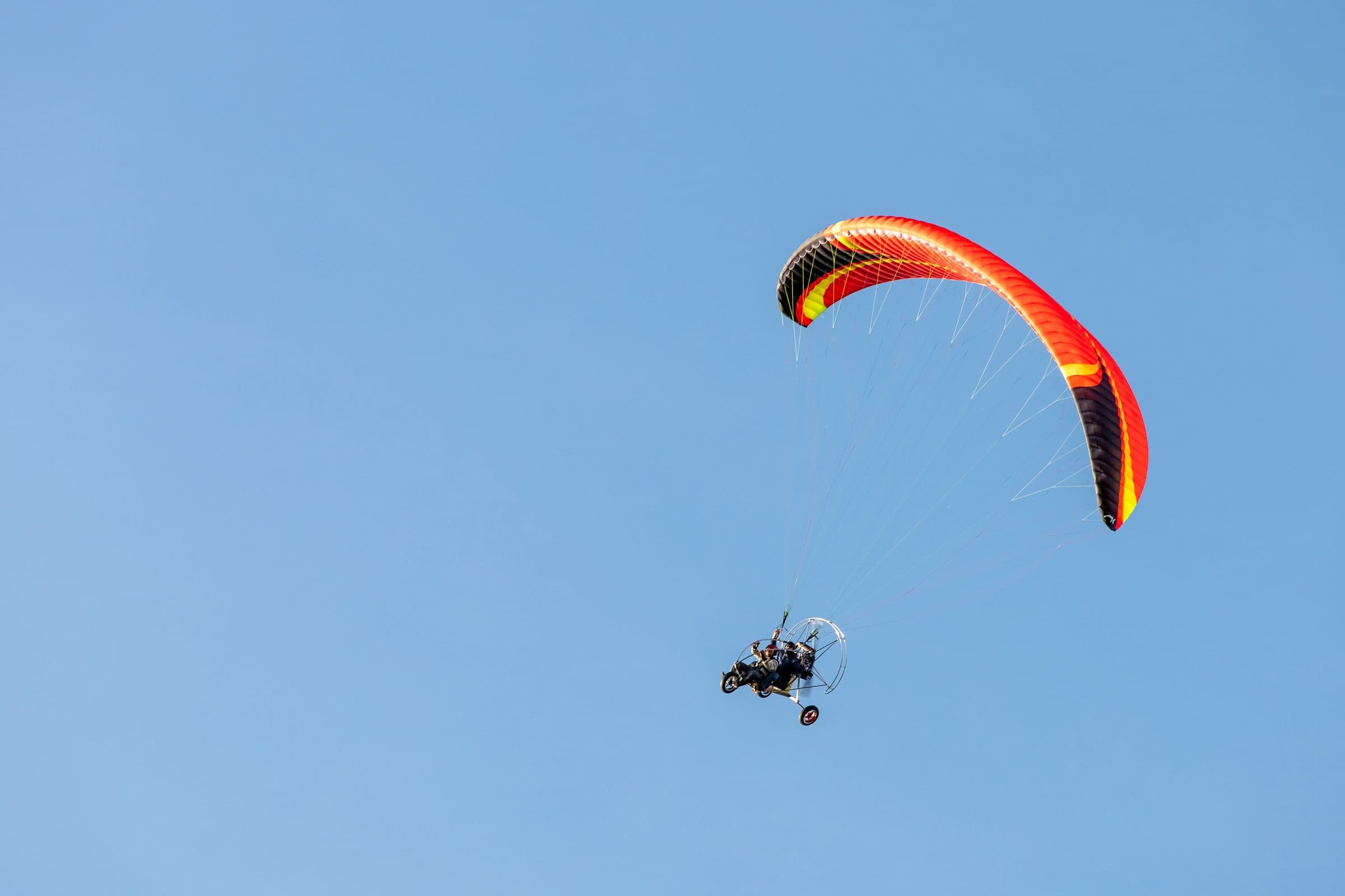 Paramotor trike flying under a bright red-orange wing in a blue sky