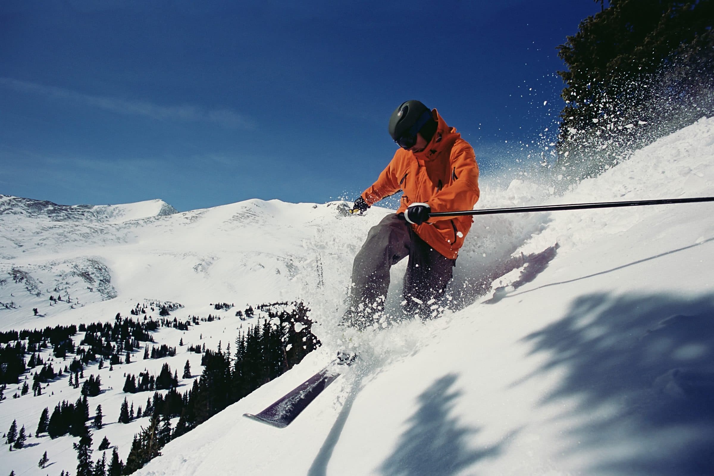 Skier in an orange jacket carving powder with spray, high alpine bowl and deep blue sky