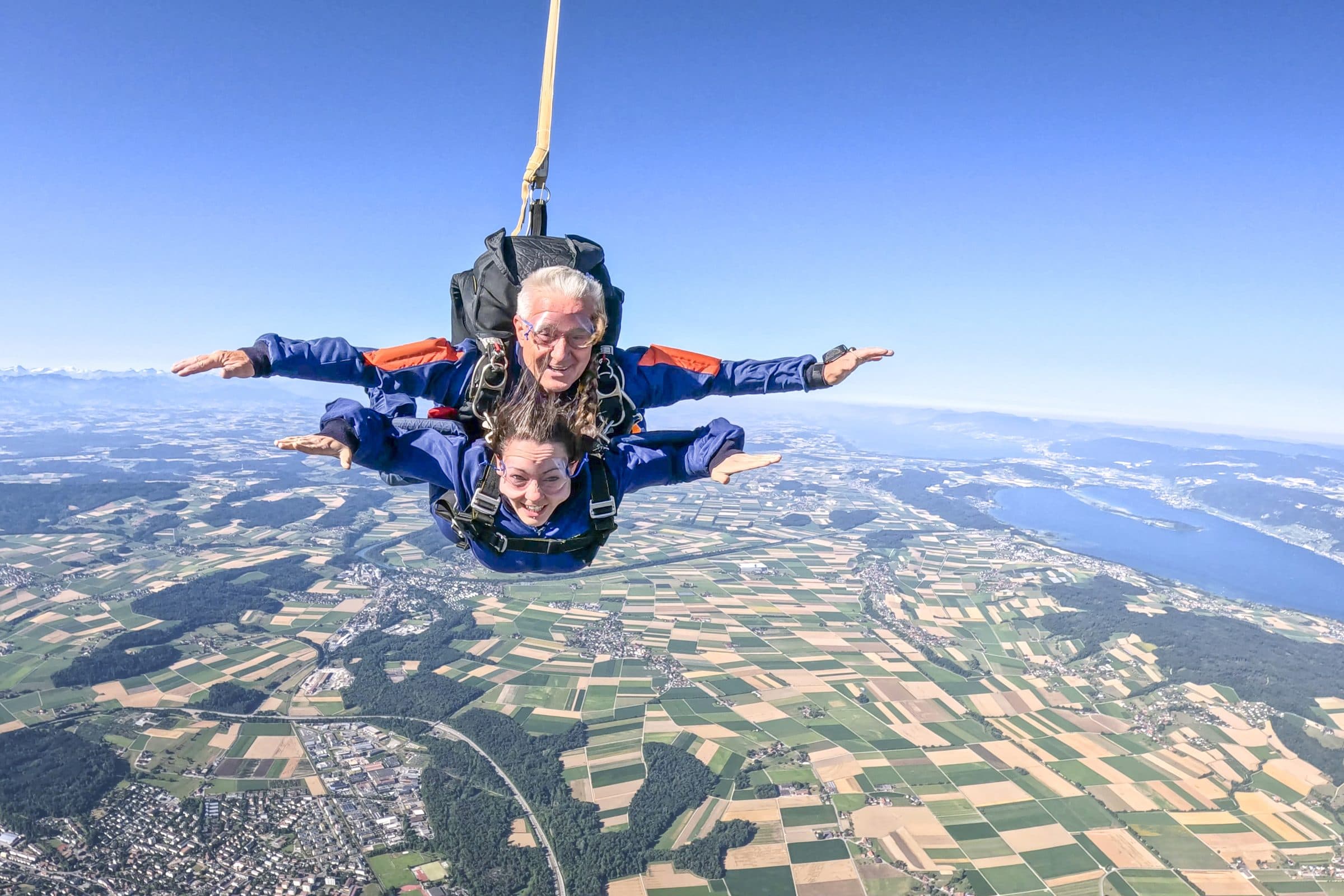 Tandem skydivers in freefall with drogue, both smiling, patchwork farmland far below