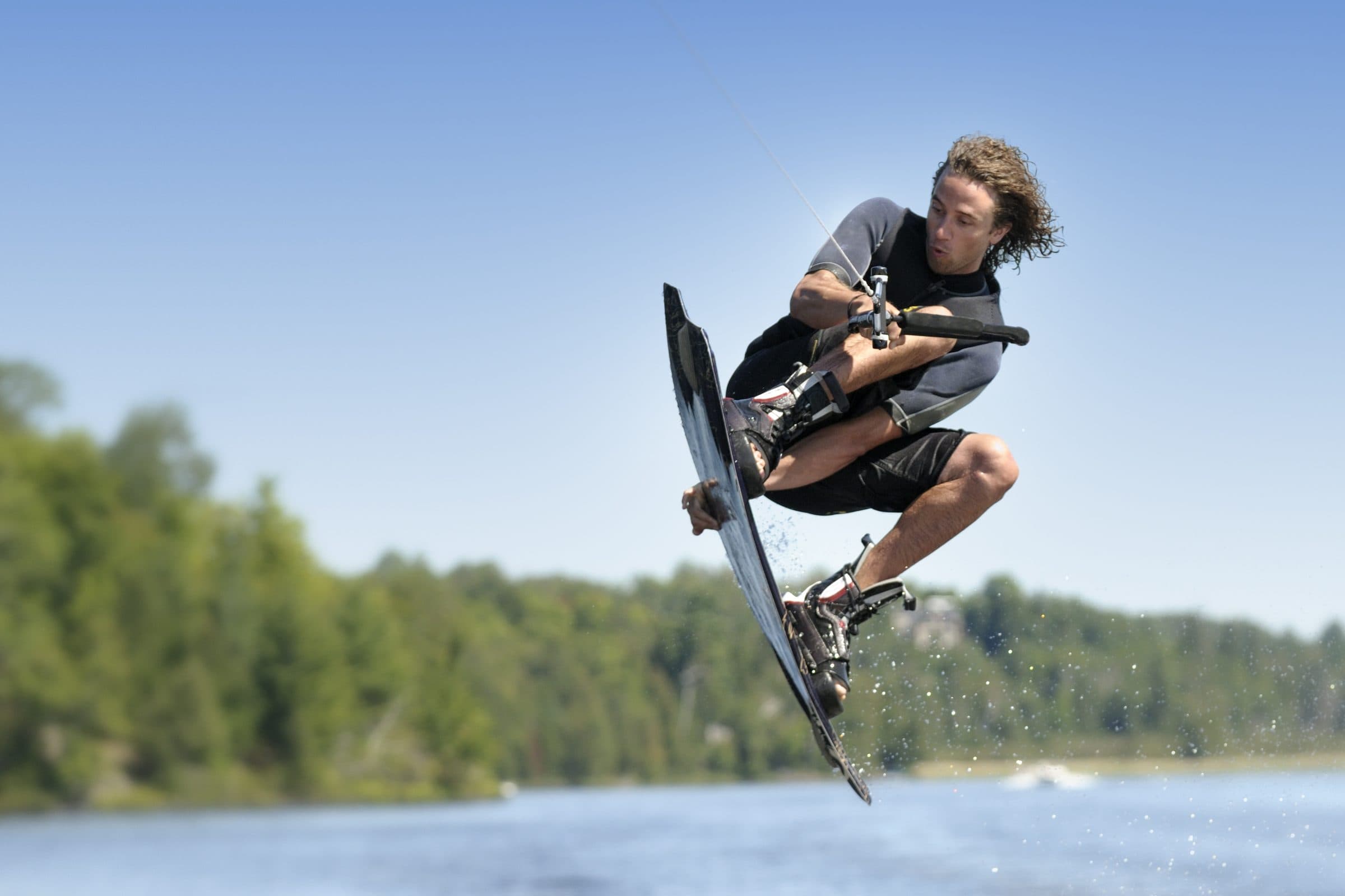 Wakeboarder airborne grabbing the board, handle in hand, blurred lake and trees behind