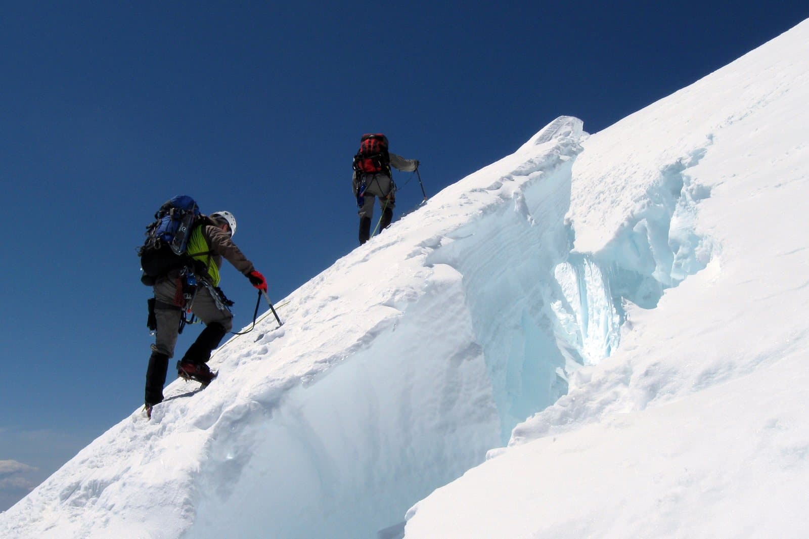Two roped mountaineers with ice axes climbing a steep snow ridge past a crevasse