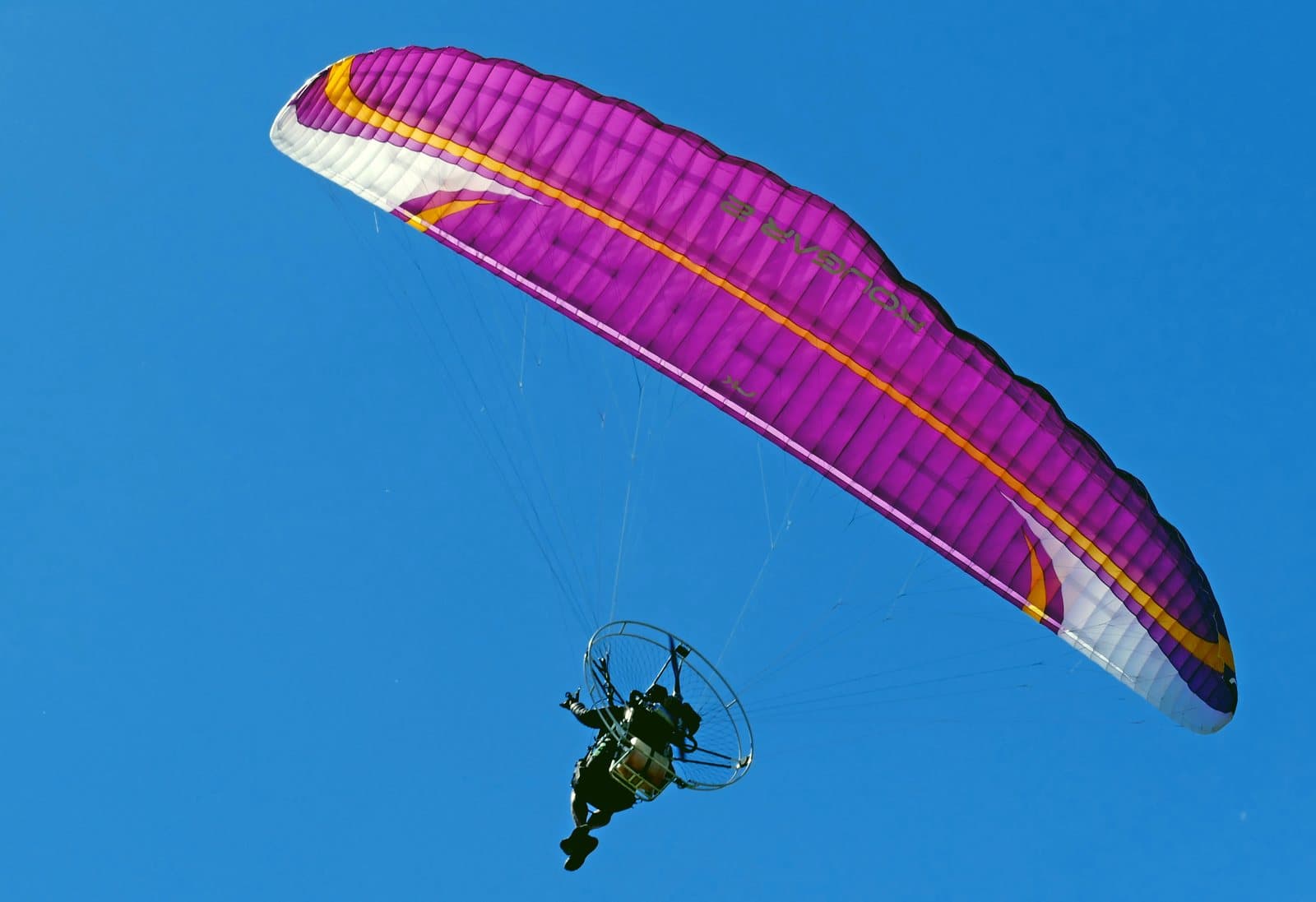 A paramotor pilot flying under a pink and white wing against a blue sky