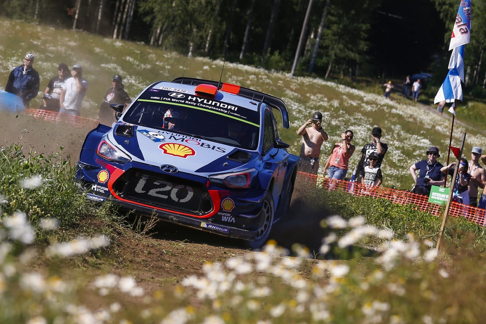 A rally car sliding on a gravel stage past spectators in the trees
