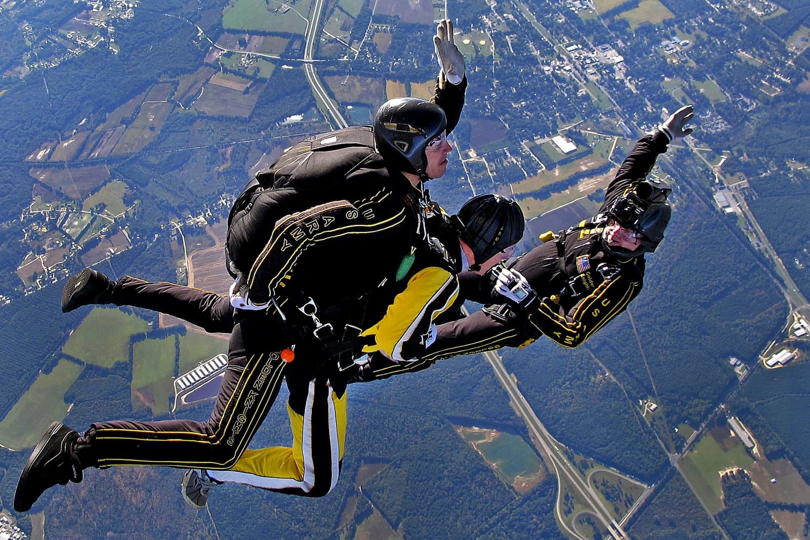 A tandem skydiving pair in freefall with a third skydiver alongside, high above farmland