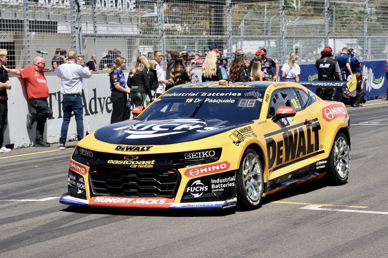 A yellow race car cornering on a street circuit with spectators behind the fence