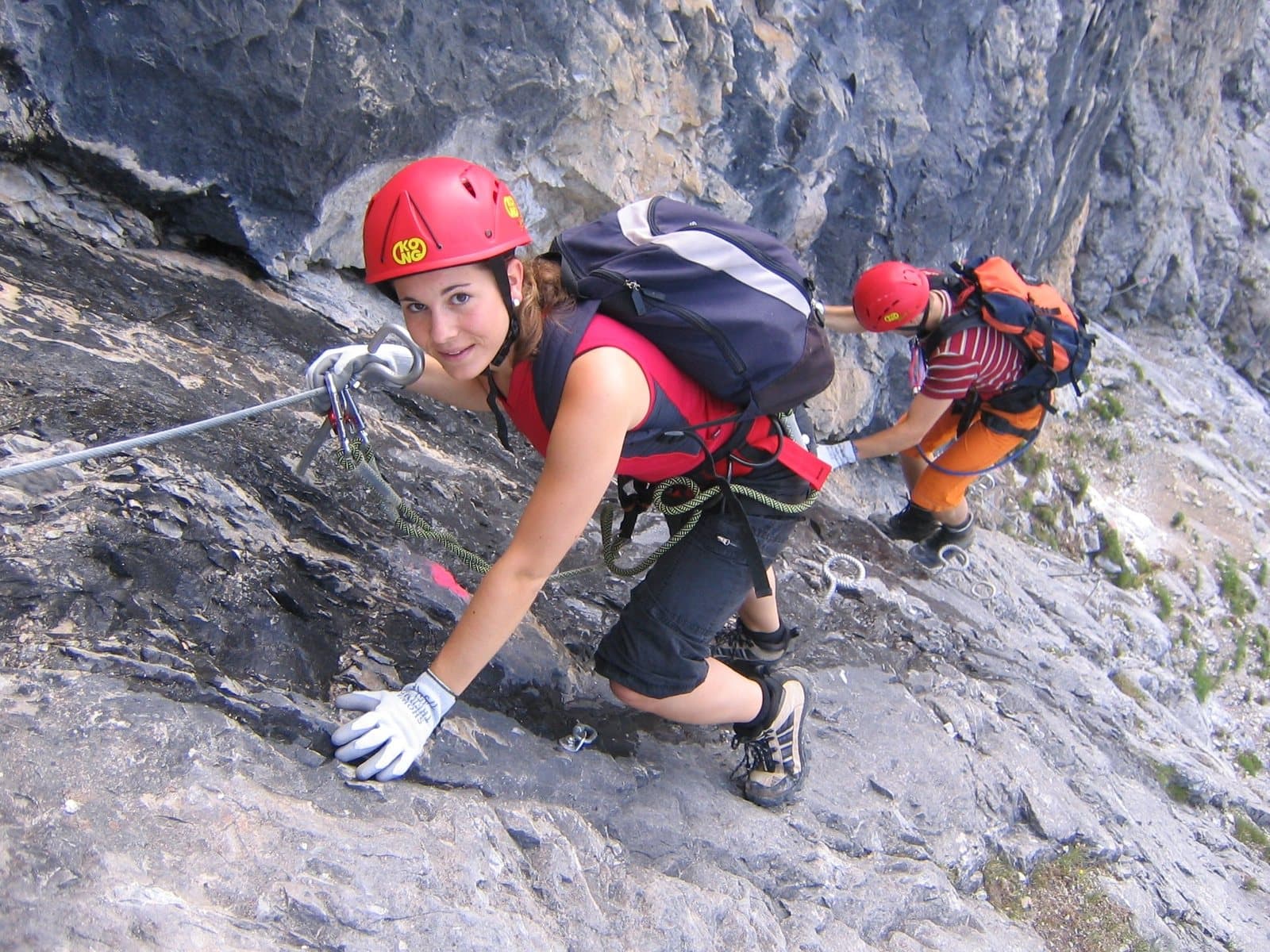 Two climbers in helmets clipped to a fixed steel cable on a steep rock face