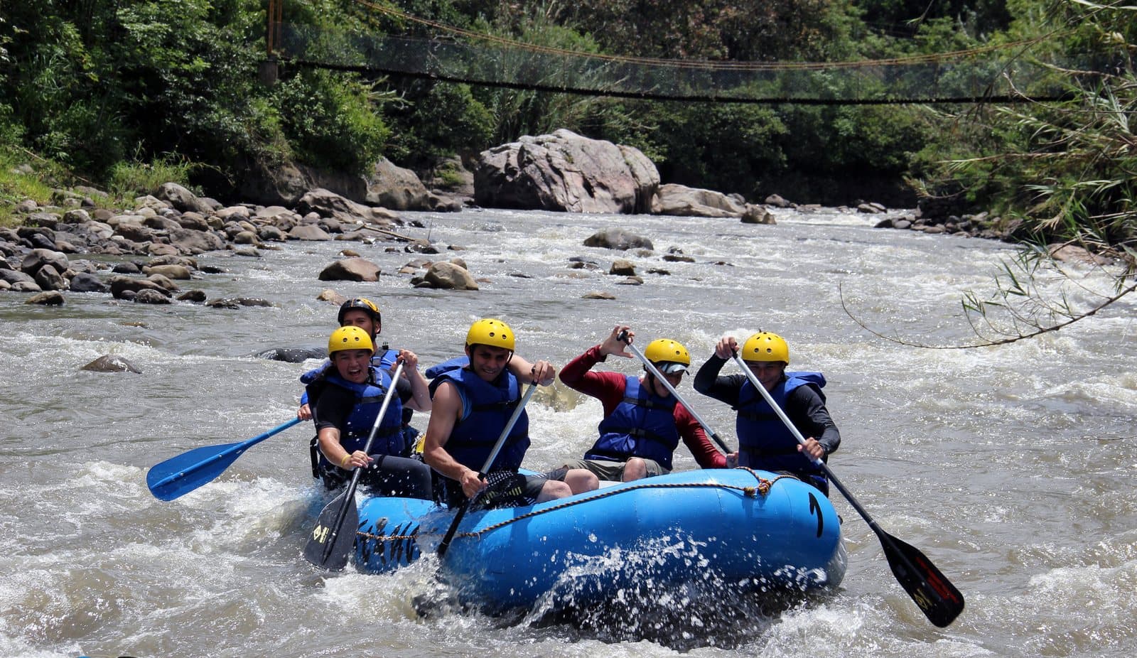 Five rafters in helmets paddling a blue raft through whitewater