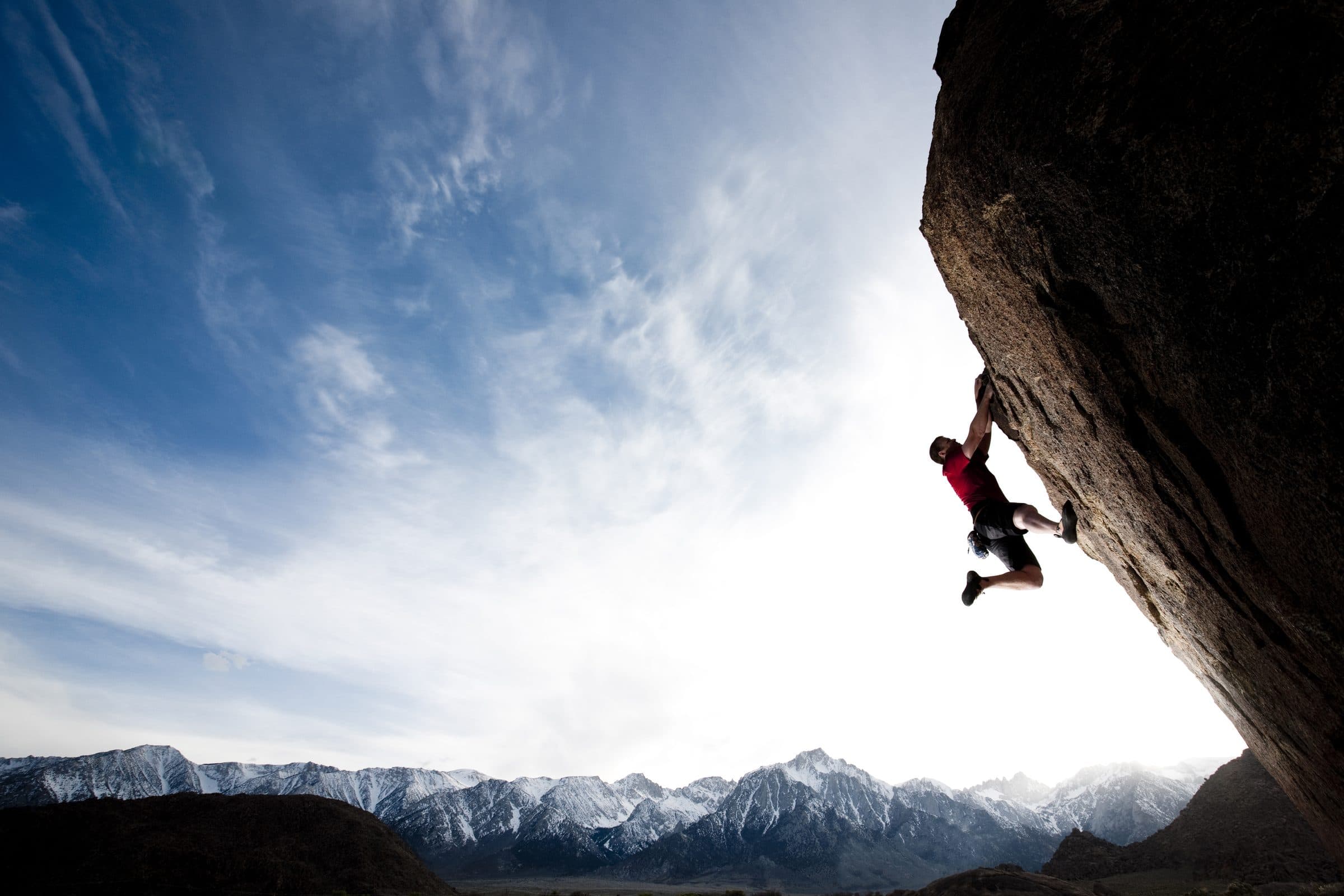 Climber on a big overhang against a bright sky, snowy mountain range below